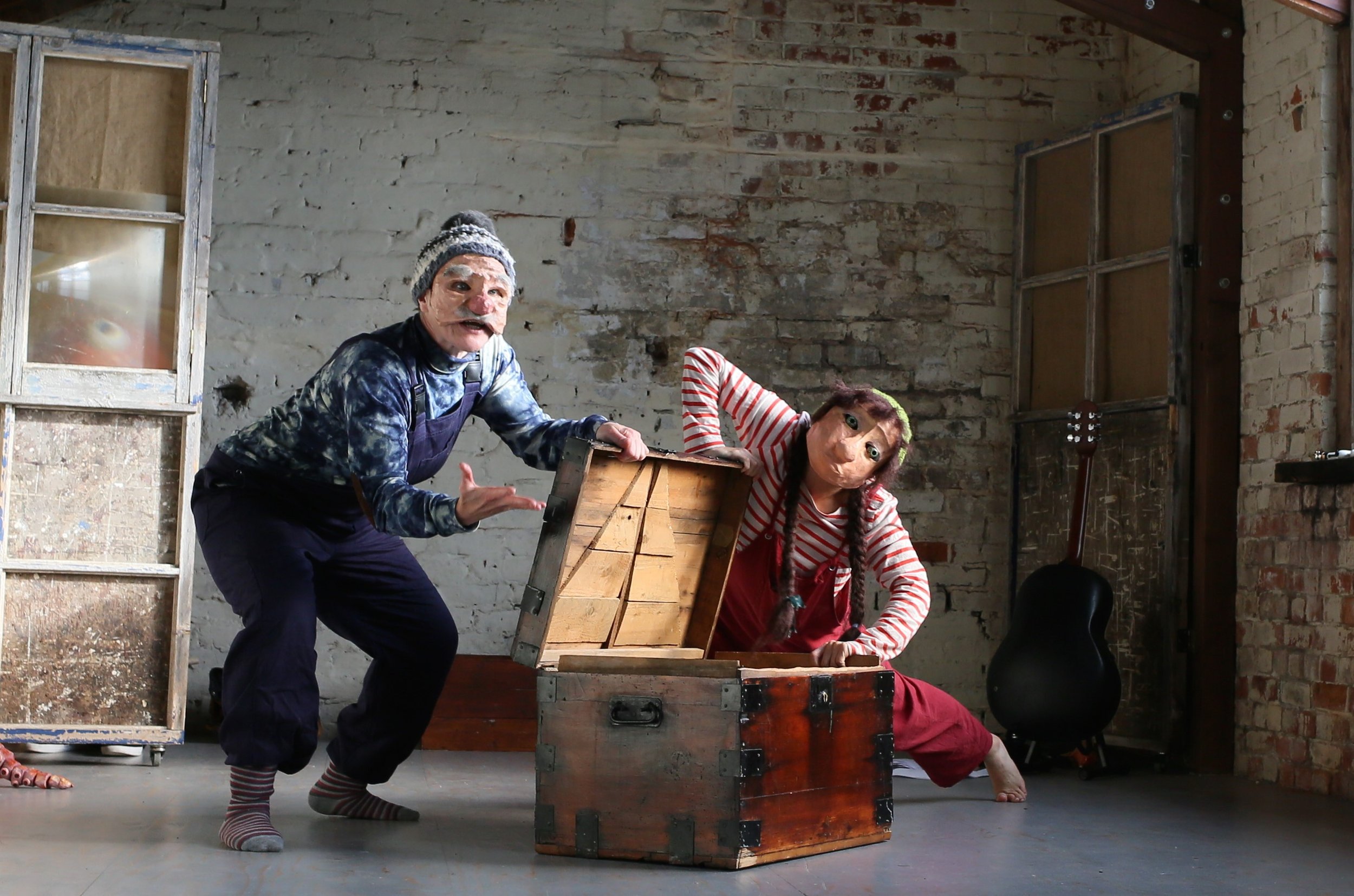 Two people wearing masks and colorful clothing play inside a room with brick walls. They are interacting around an open wooden chest, with one person in a striped shirt leaning inside and the other gesturing.