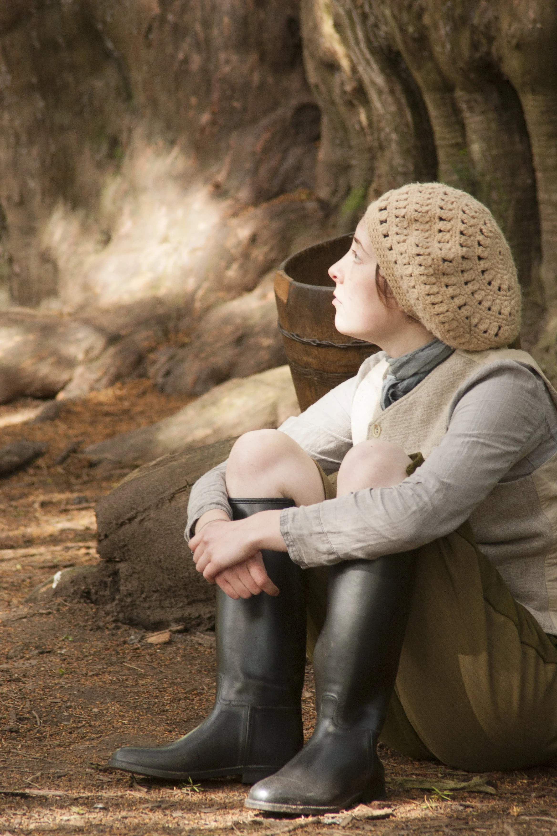 A woman wearing a beige knitted hat, beige shirt, and black rubber boots, sitting outdoors in a forest, gazing up at the trees.
