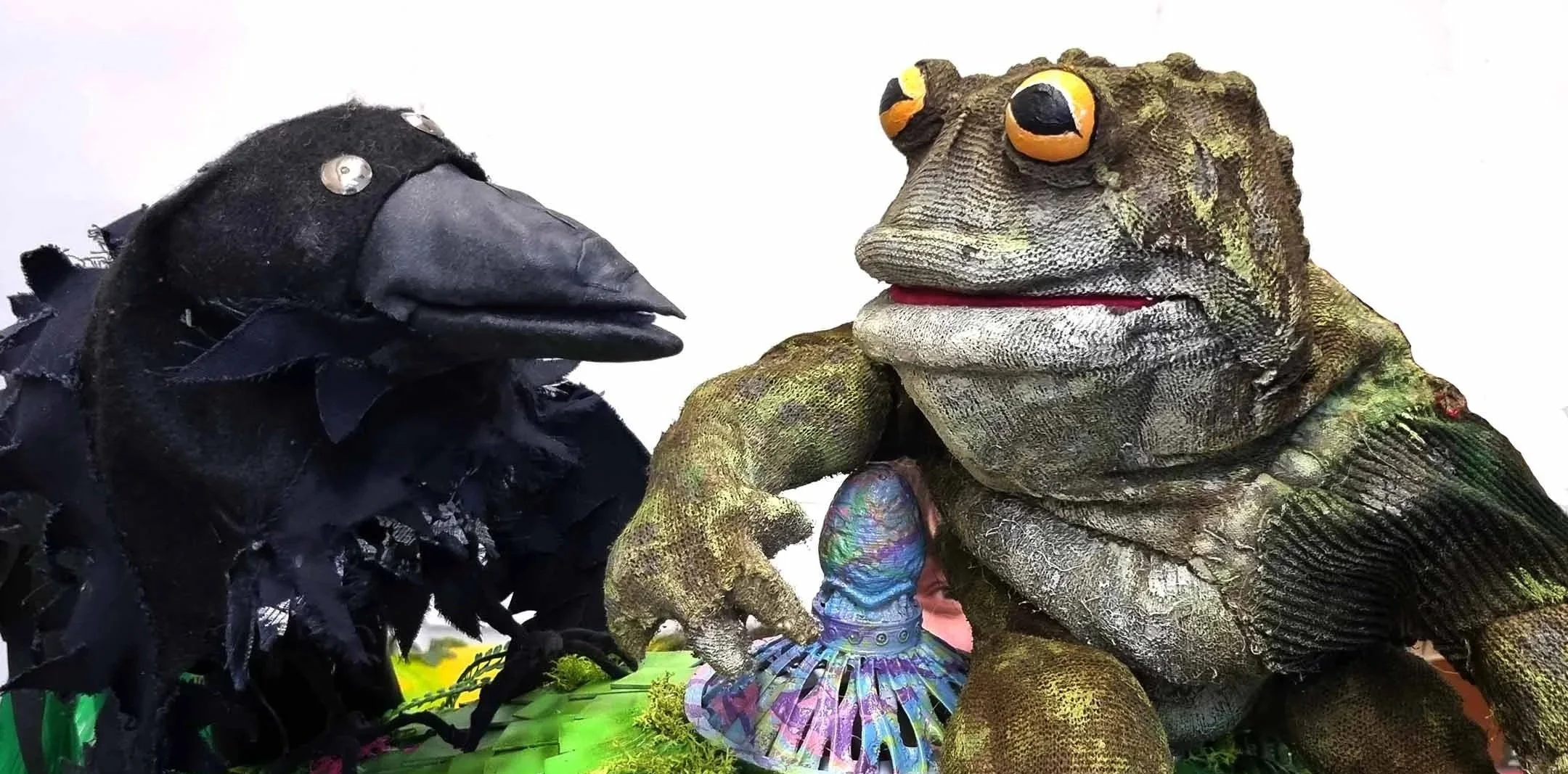 Two puppet animals, a black crow and a green toad, sit on a colourful display with green grass and purple decoration, against a plain white background.