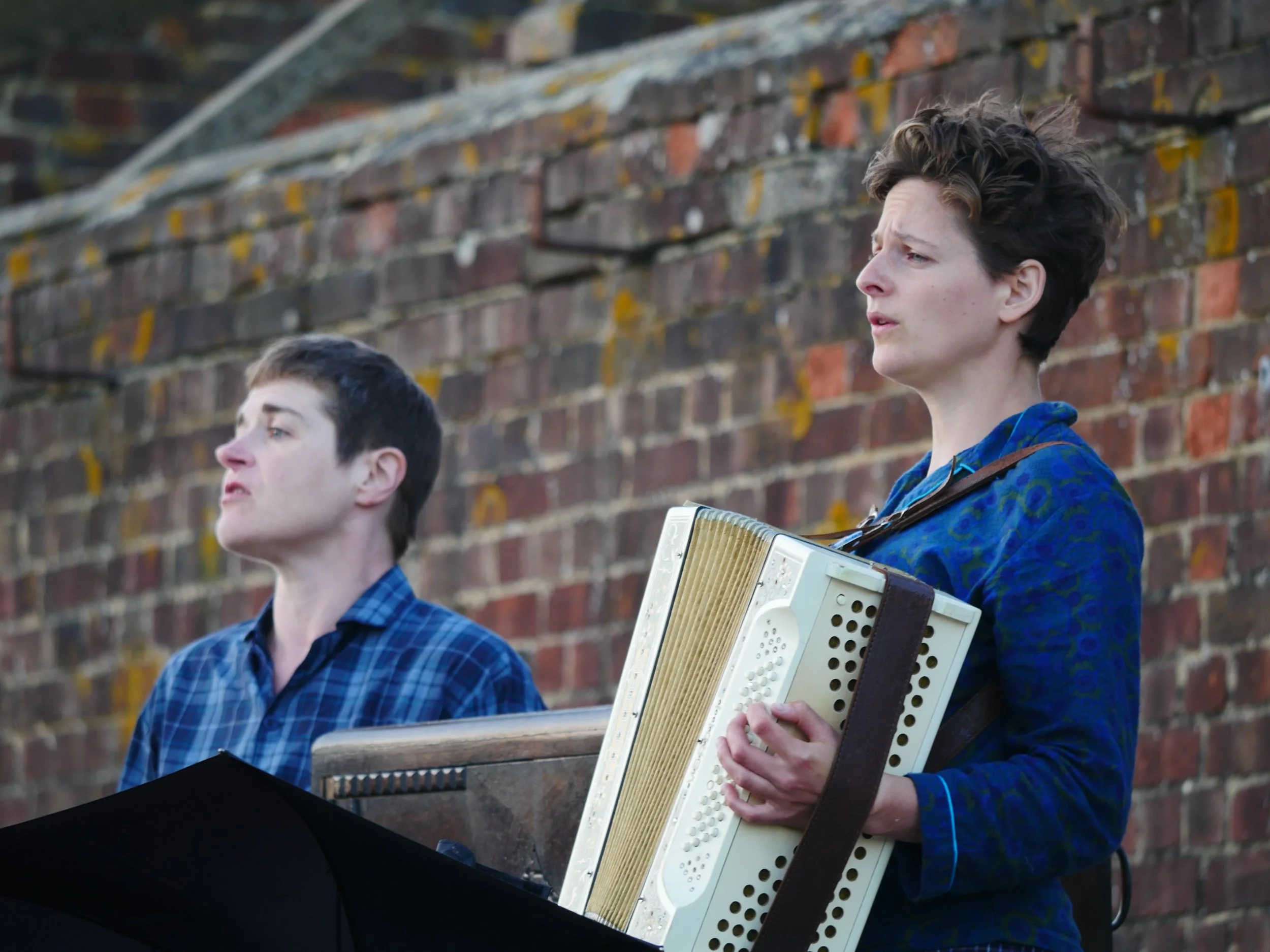 Young man with short curly hair and light skin playing an accordion, standing outdoors against a brick wall.