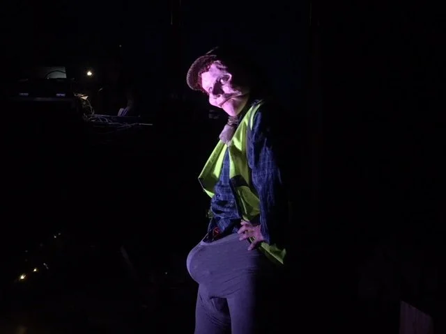 A person wearing a high-visibility vest, a gray hoodie, and gray pants, standing with hands on hips in a dark environment, illuminated by purple lighting.
