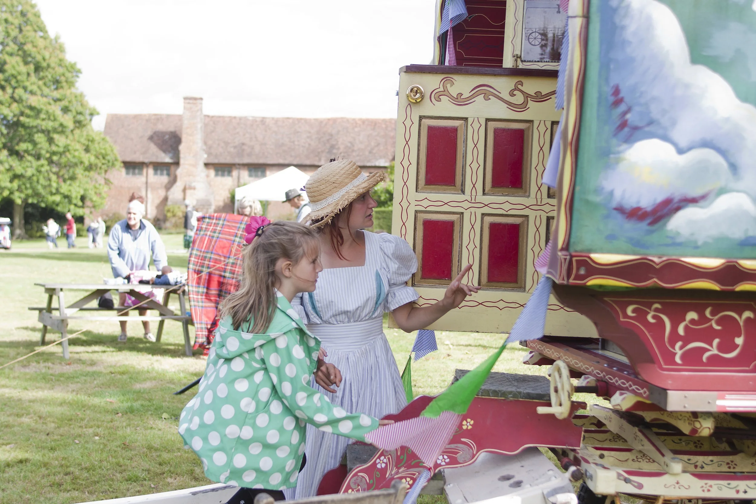 A woman and a girl are at a carnival game booth with painted wood panels, and the woman is pointing at something. The girl is wearing a green polka-dot raincoat with a pink hair tie, and the woman is wearing a white dress with gray stripes and a stra