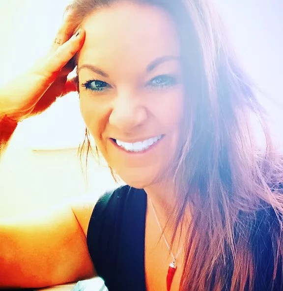 A woman with long reddish-brown hair smiling, resting her hand on her forehead, with sunlight in the background.