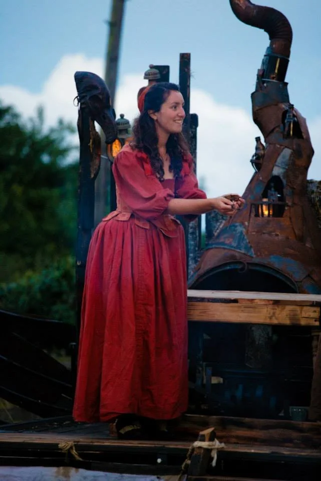 A woman in a long red Victorian-style dress smiling and holding a small object, standing next to a steampunk-inspired mechanical structure outdoors.