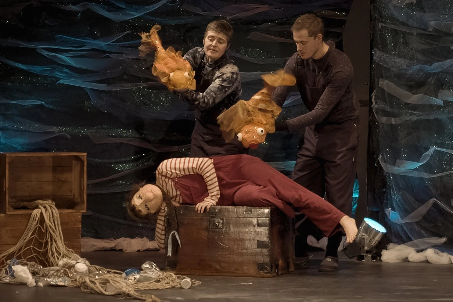 People performing with puppets that have large eyes and orange hair, one puppet lying in a chest and others being manipulated, on a stage with a dark, textured backdrop, scattered ropes and props.