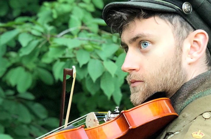 A man wearing a military uniform and cap is playing a violin outdoors, with green foliage in the background.