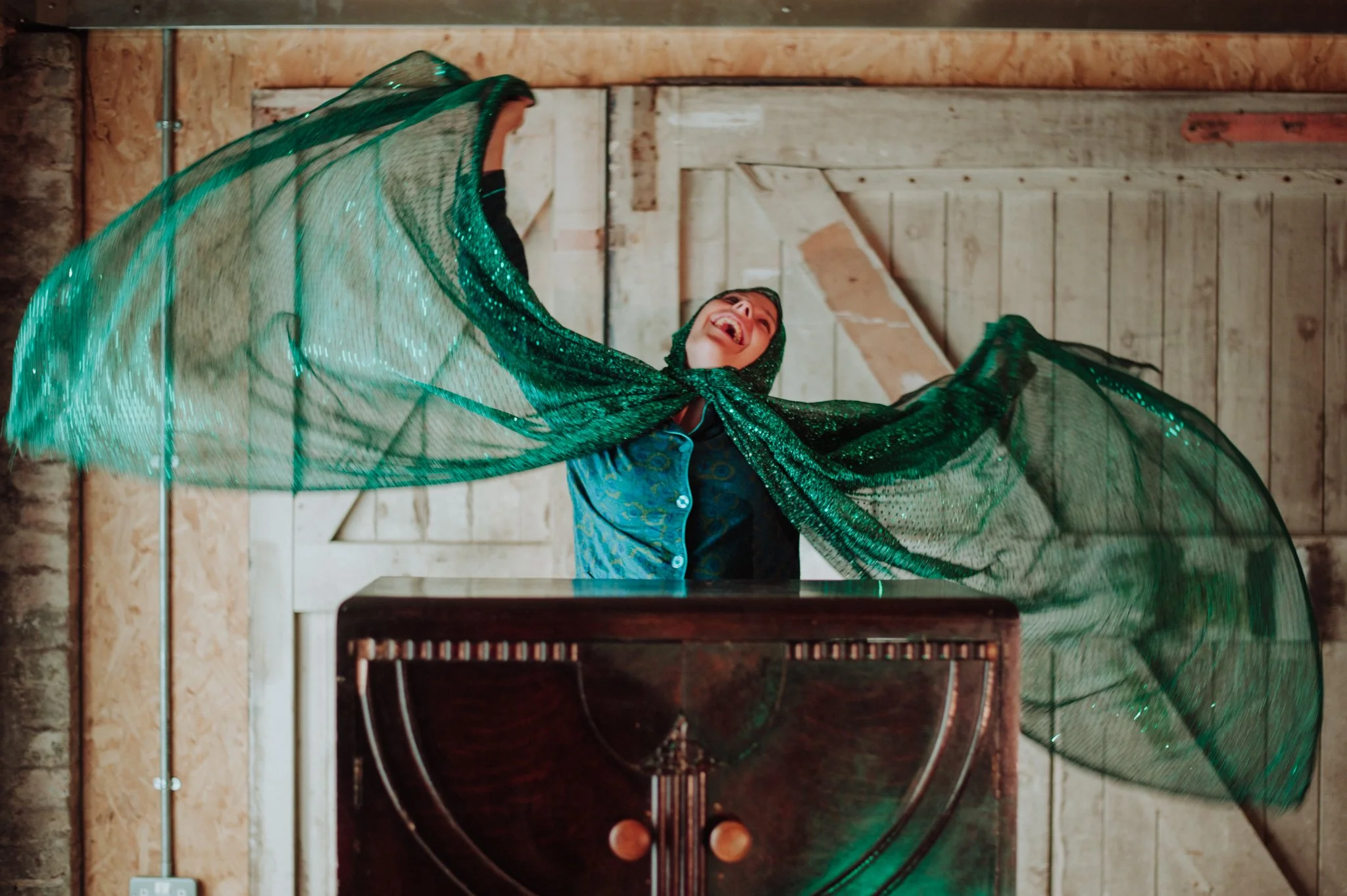 Woman smiling and looking up, wearing a sparkling green shawl and a dark outfit, in a rustic wooden room.