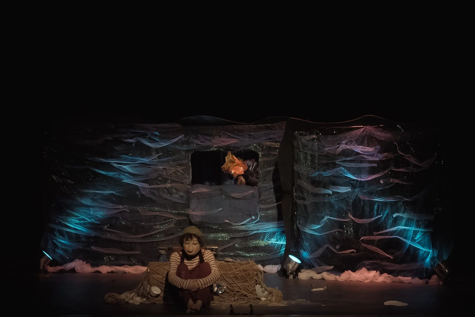 The photo shows a theatrical stage scene with two characters, one sitting on the ground in front, wearing a striped sweater and a mask, and the other standing in a window or opening of a large, abstract backdrop made of layered fabric and lights. The