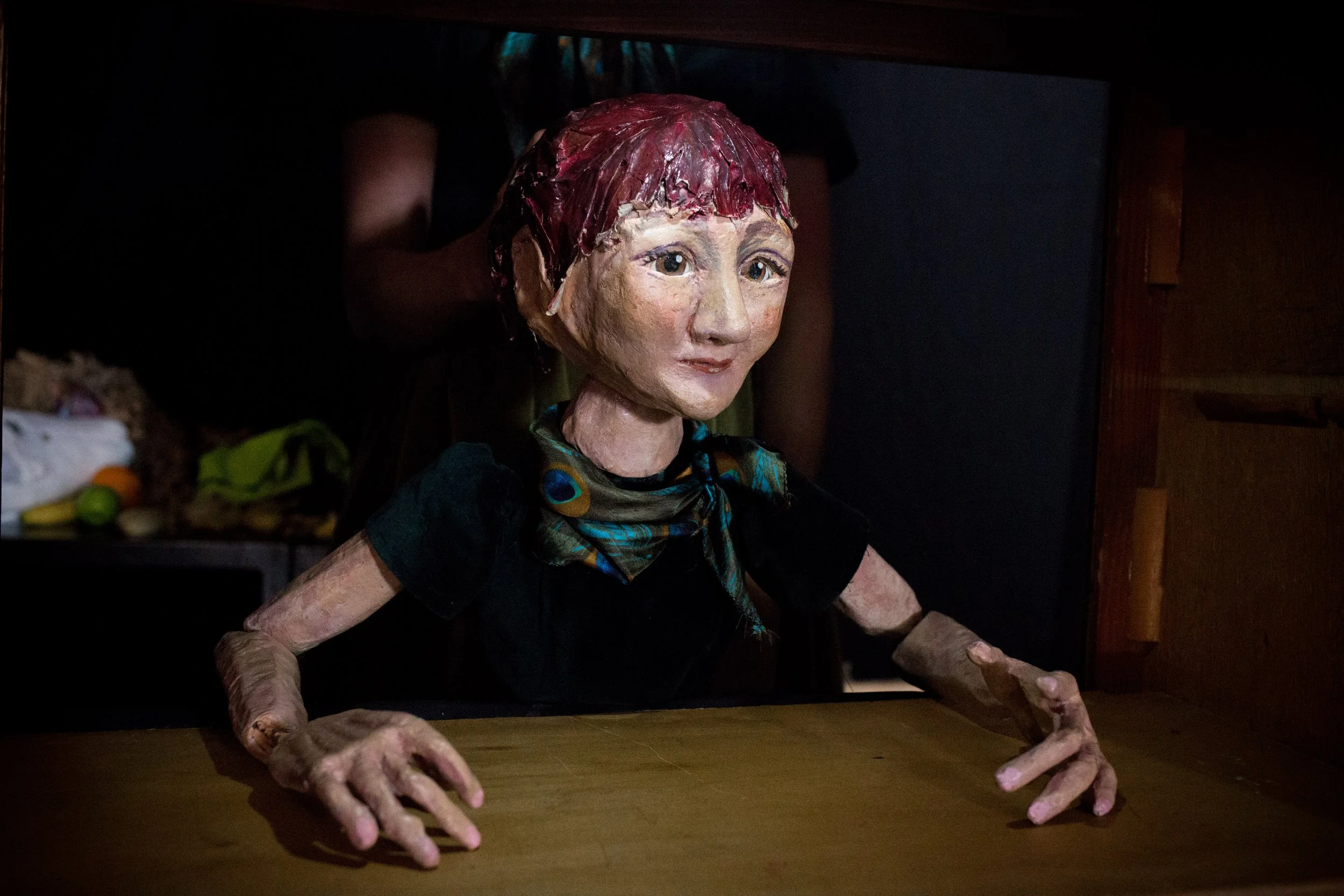 A detailed puppet of a woman with red hair, painted face, wearing a colorful scarf, leaning over a wooden table with hands extended on the surface, in a dimly lit room.