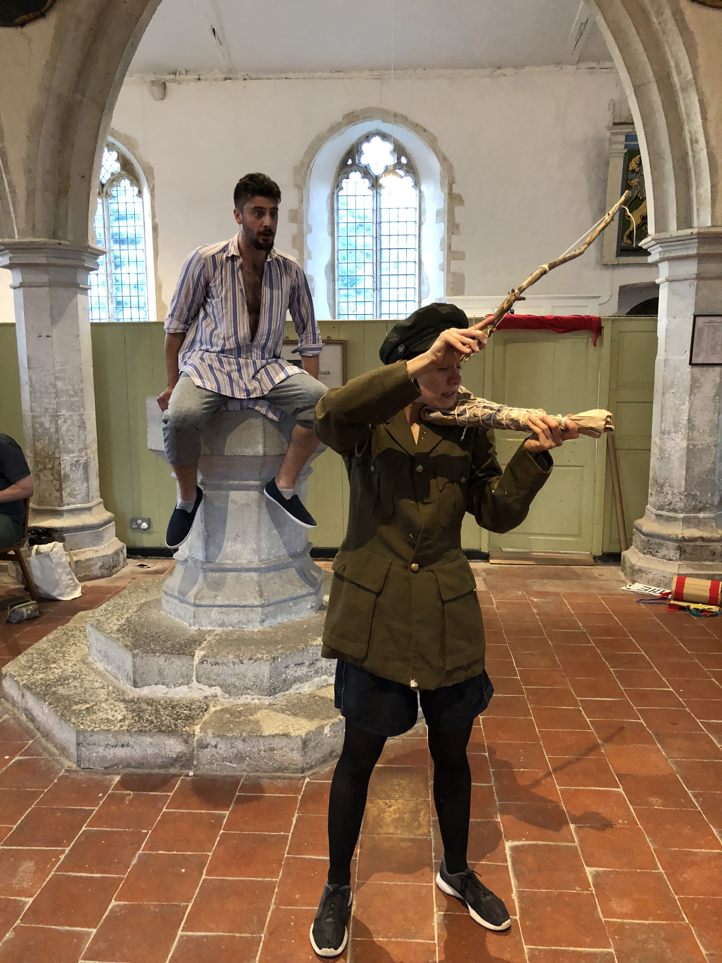 A person dressed as a soldier holding a bow and arrow inside a church, with another person sitting on a stone pillar and a man sitting on the ledge with a surprised expression.