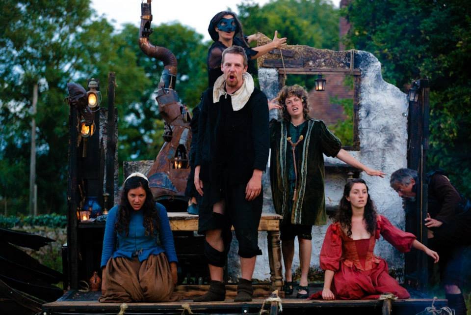A theatrical outdoor performance with five actors on a rustic stage, dressed in medieval or fantasy costumes, surrounded by greenery and set pieces resembling a whimsical village scene.