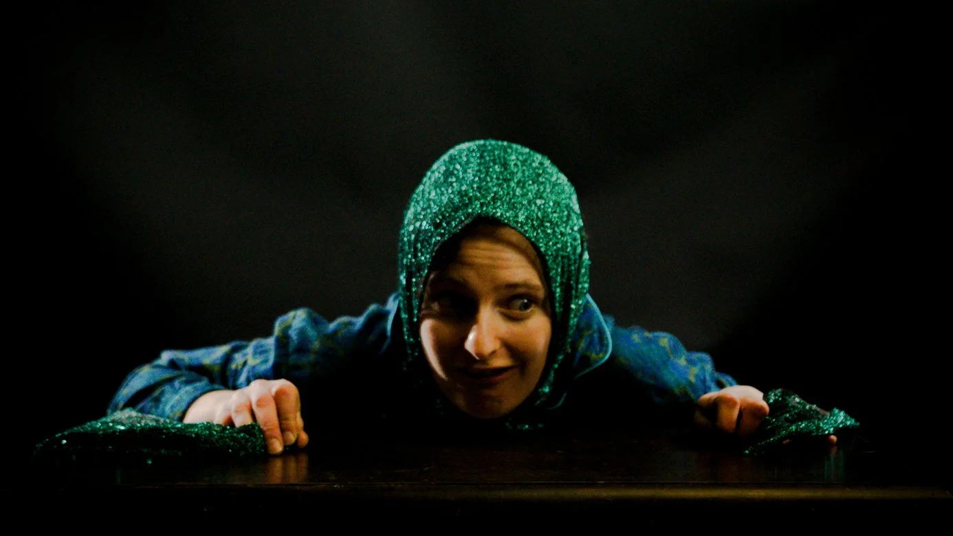 Person wearing a shiny green headcovering and blue clothing with a playful expression, leaning on a dark surface against a black background.
