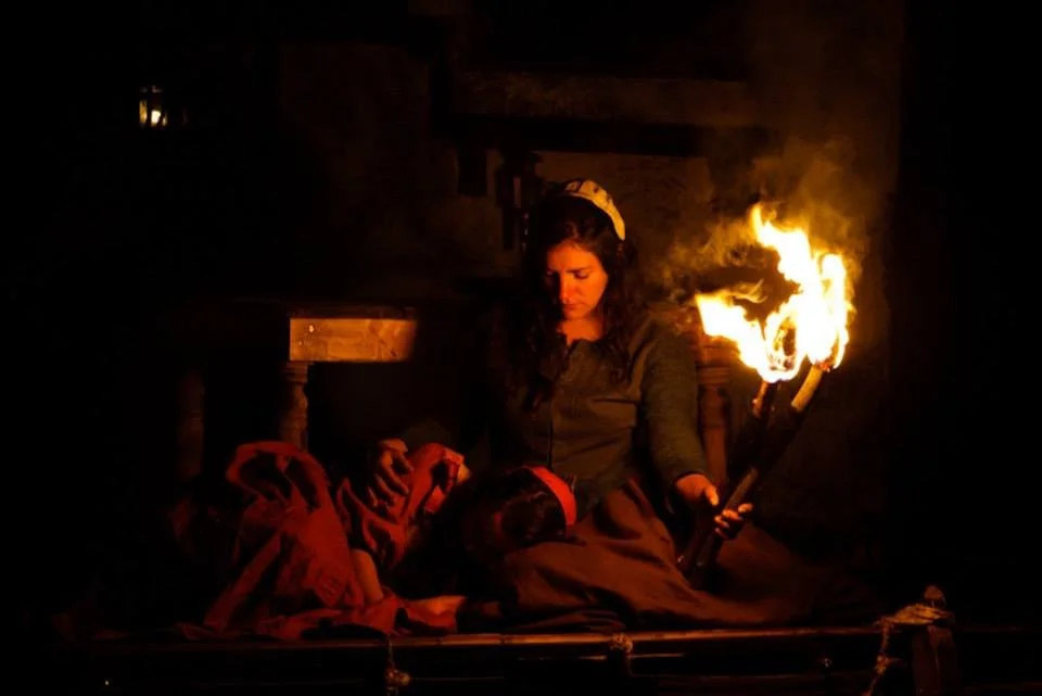 A woman sitting in a dark room with a fire torch in her hand and a helmet on her head, illuminated by the fire's glow.
