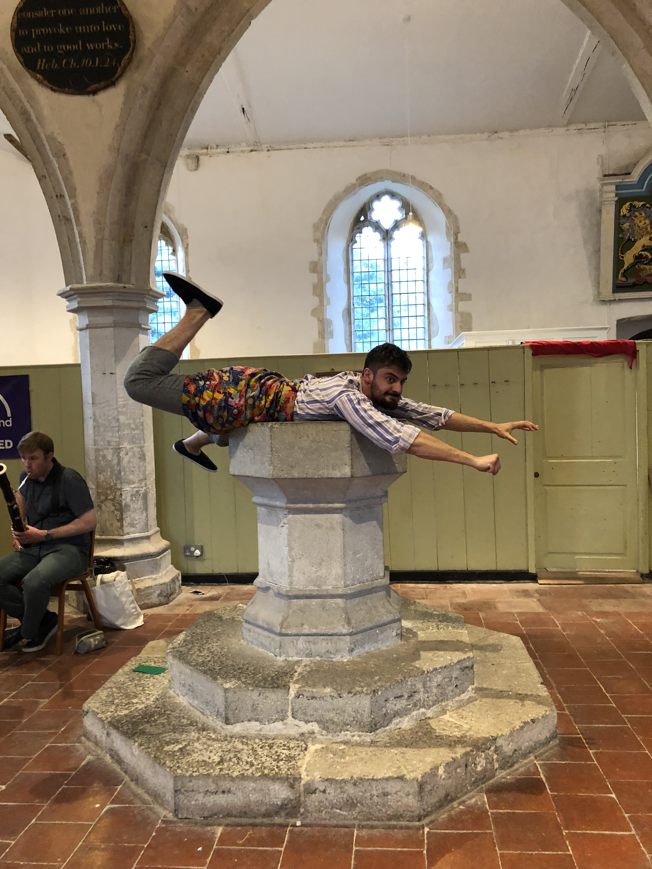 A man is lying on top of a church baptismal font with his arms and legs extended outward. The baptismal font is made of stone with multiple tiers, situated on a stone base, inside a church with arched windows and historic architecture.