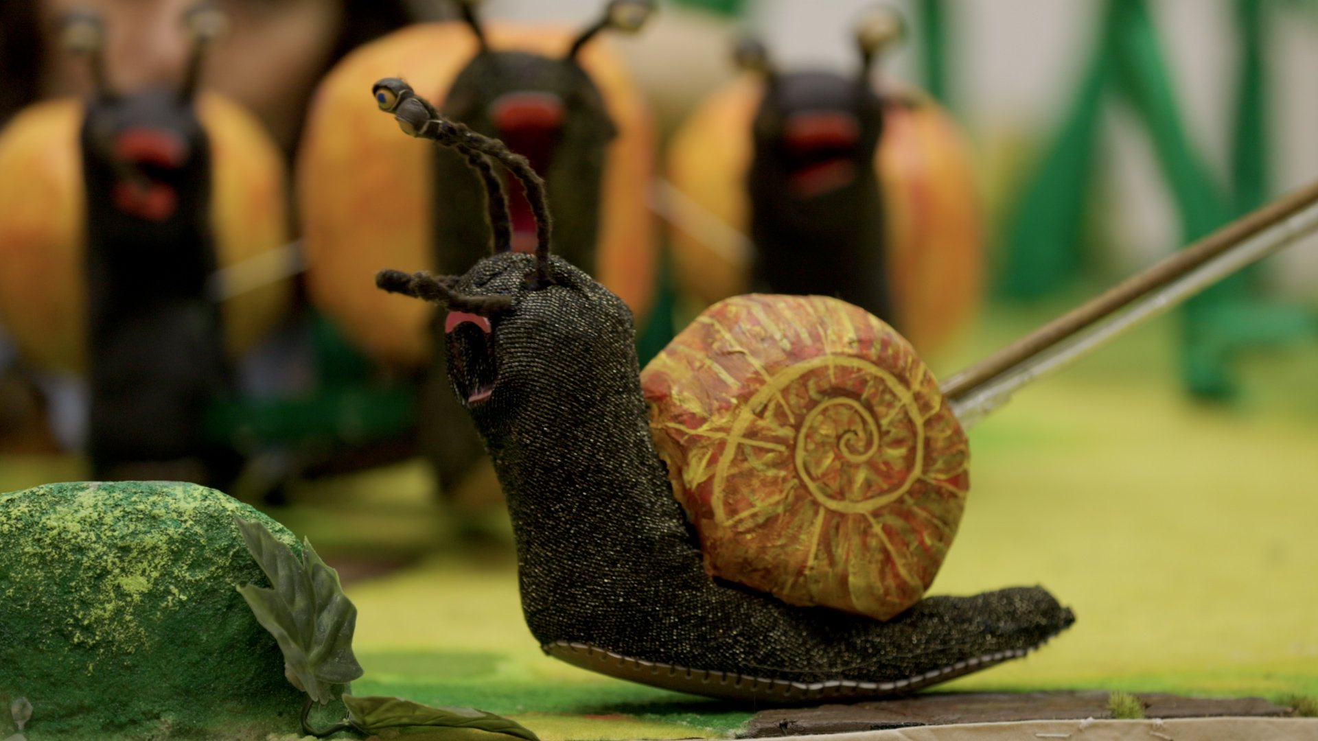 A puppet snail with an orange and yellow shell, and yellow antennae, with a backing chorus of puppet snails, surrounded by other puppet plants.