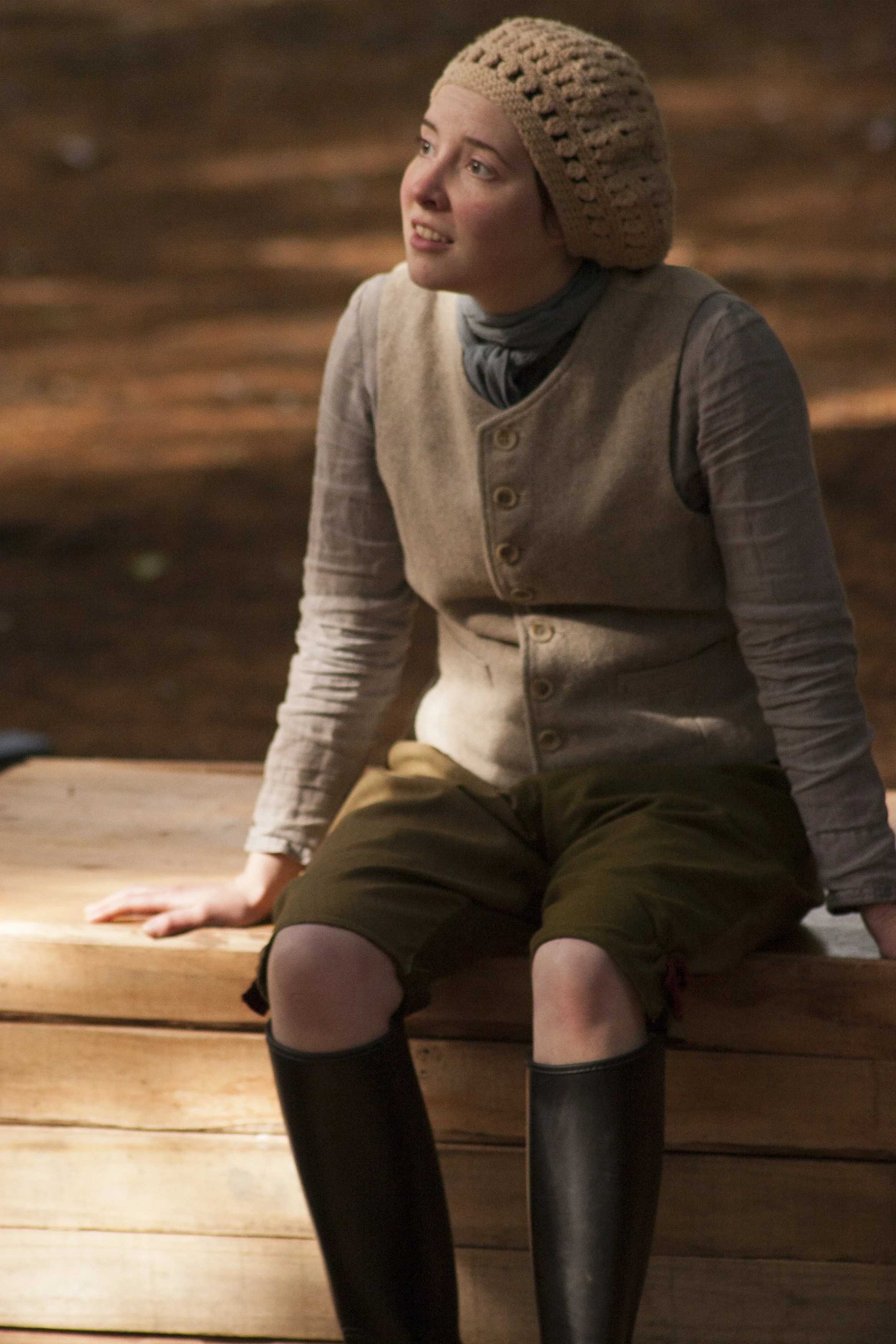 A woman sitting on a wooden surface, wearing a grey vest, brown skirt, black knee-high boots, and a beige knit hat, looking slightly upwards.