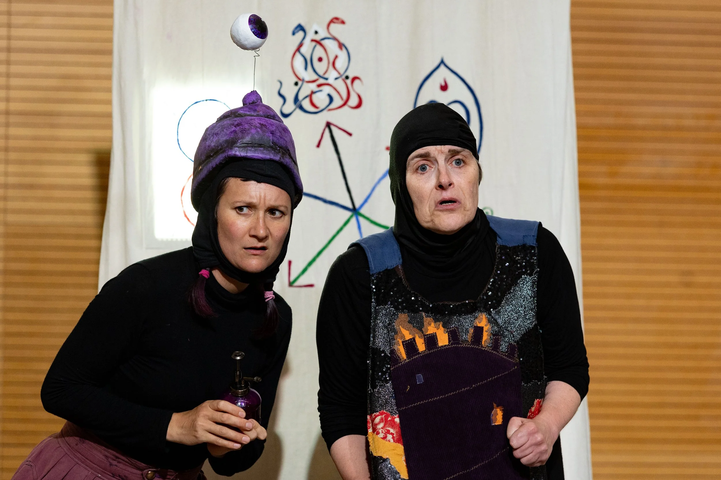 Two women dressed in dark clothing with head coverings, standing indoors in front of a white curtain with a colorful abstract design. One woman holds a purple balloon with an eyeball decoration attached by a string.