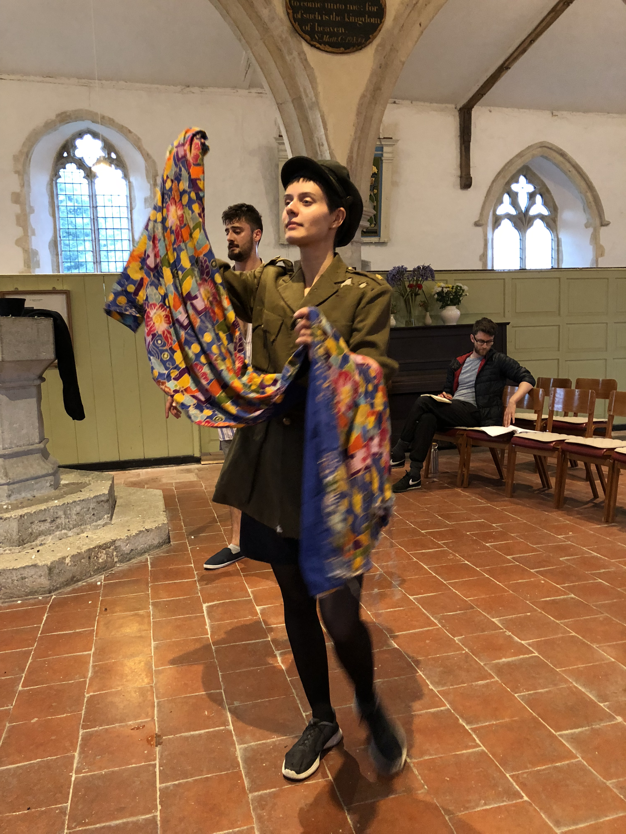 A woman in a military-style uniform is holding a colorful, patterned fabric while standing inside a church with stained glass windows and wooden pews. A man and another person sit in the background.