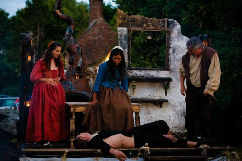 A theatrical outdoor scene with four actors, two women and one man, surrounding a man lying face-up on the ground, appearing to be part of a performance concerning a historical or fantasy setting.