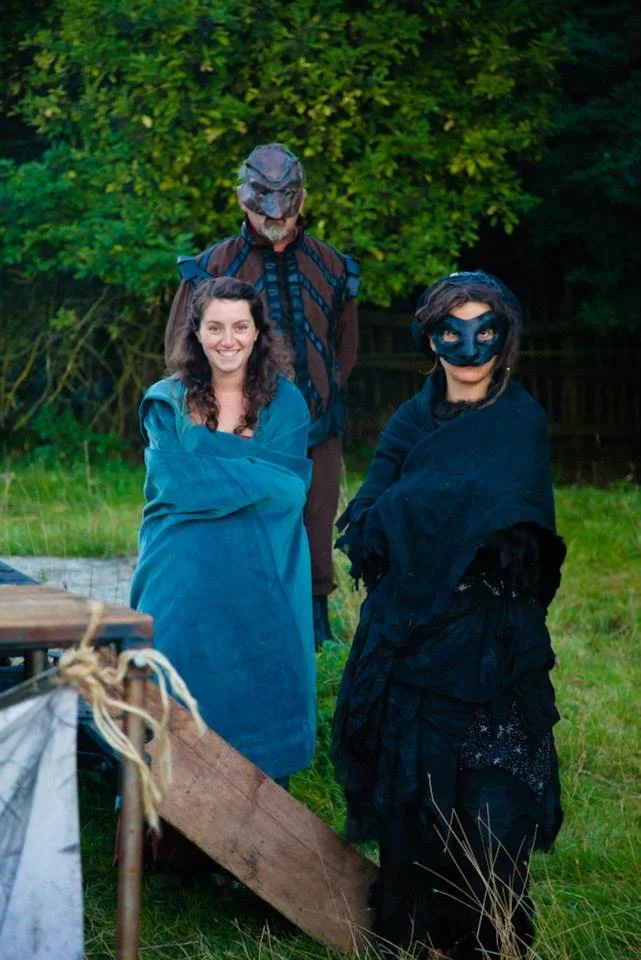 Three people outdoors in costume at night, two women dressed as characters from Robin Hood, with face masks and capes, and a man dressed as a dinosaur with a mask and carrying a backpack.