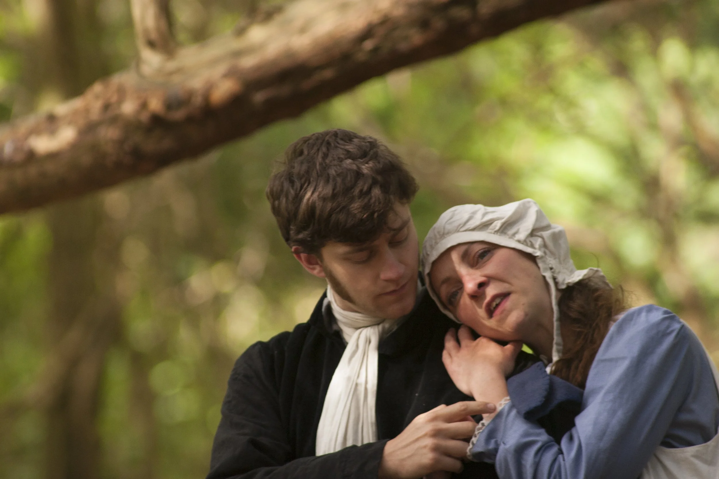Two people, a young man and a young woman, are embracing and smiling in a forest setting with green leaves and a tree branch in the background.