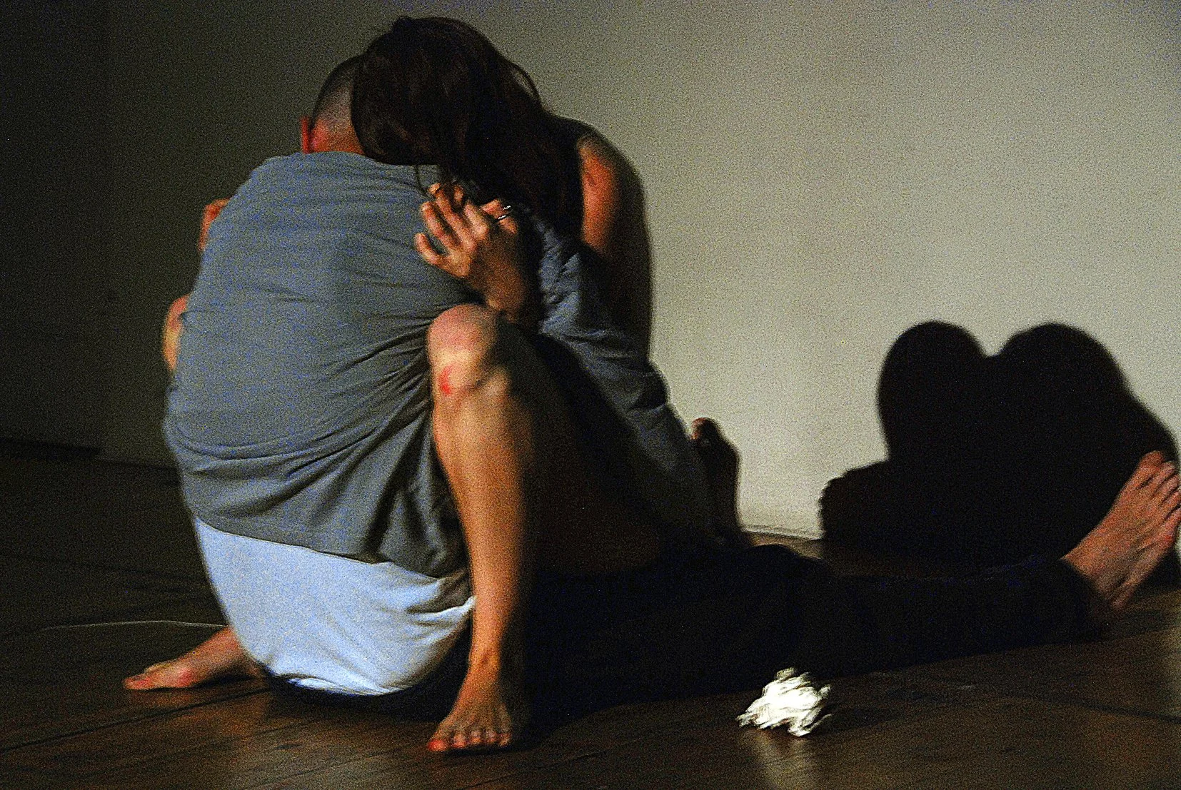 A man and a woman engaged in an intimate embrace on a dark wooden floor, with the man holding the woman close, their faces near each other, and the woman wearing dark clothing, while the man is dressed in lighter clothing. They are in a dimly lit roo