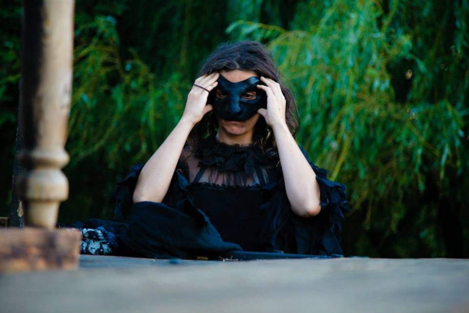 A person wearing a black mask and black dress, adjusting the mask with both hands, outdoors with green foliage in the background.