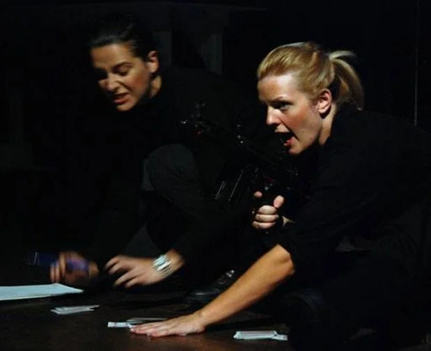 Two women are engaged in a dramatic scene, one holding a gun and the other reaching out with her hand on a table.