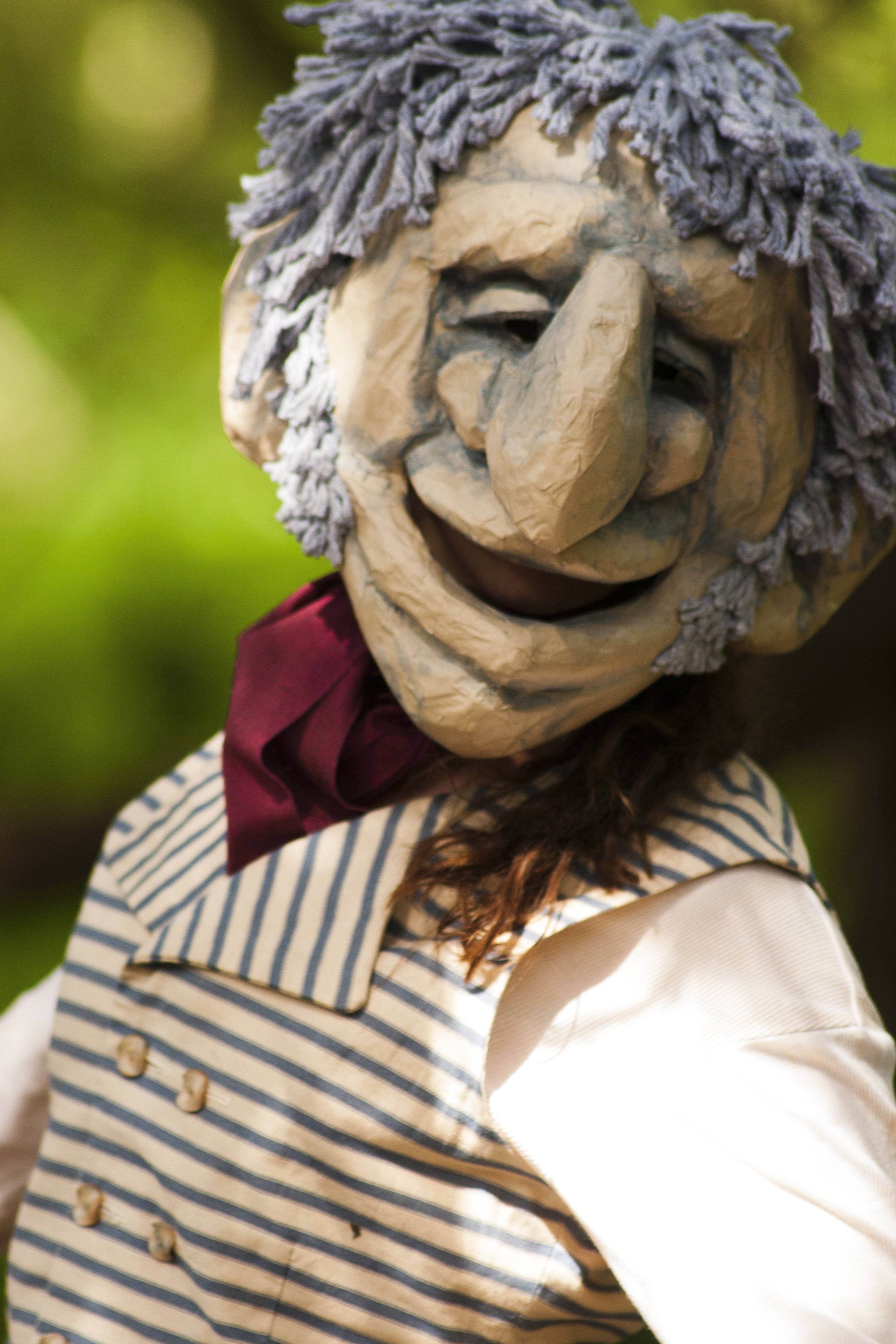 A puppet with a carved wooden face, yarn hair, and striped clothing, staged outdoors.