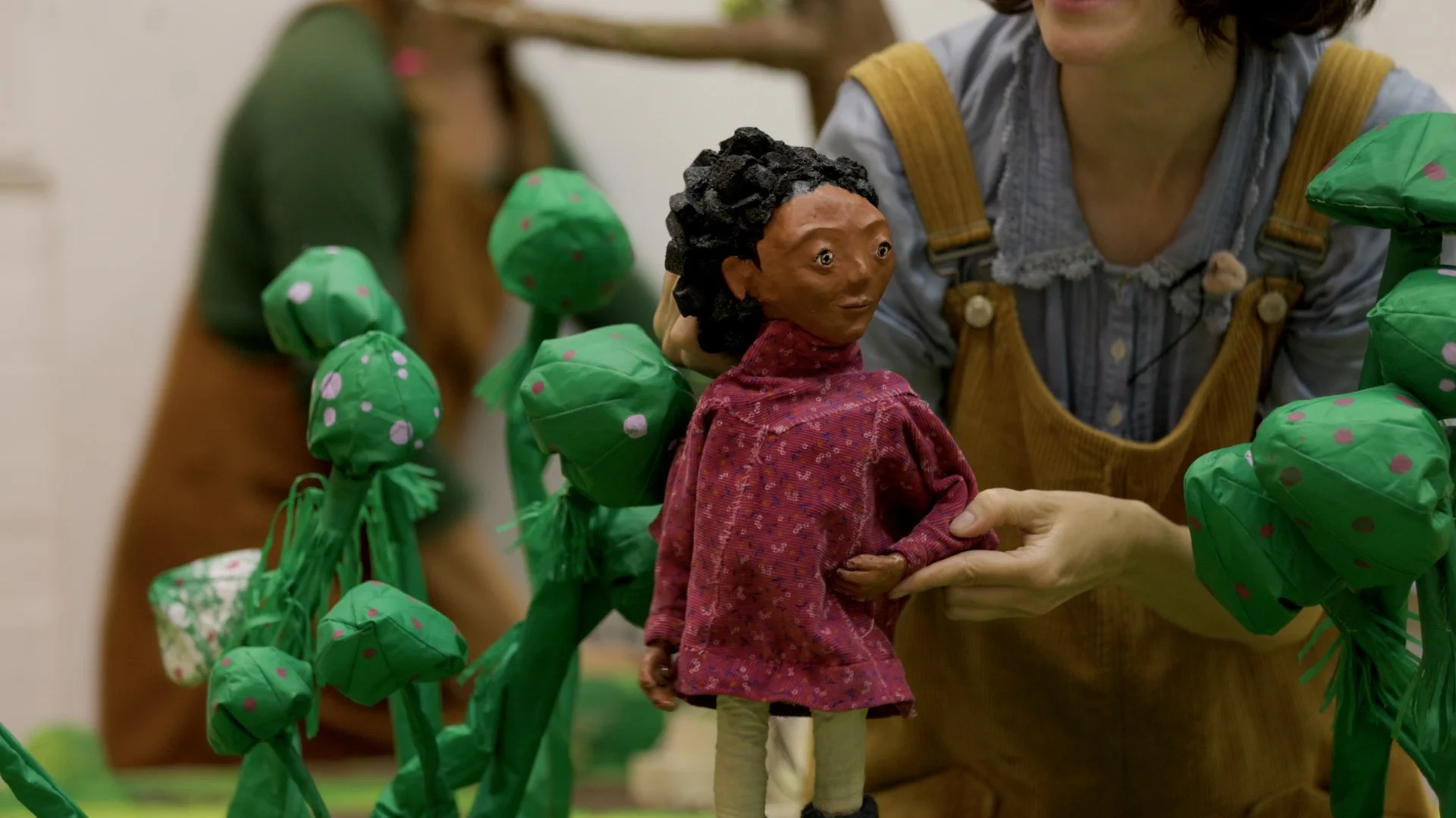 A puppet show scene with a puppet of a woman with dark curly hair, wearing a red patterned dress, being held by a person. The scene is decorated with green, paper-crafted trees with pink dots, possibly representing a garden or outdoor setting.