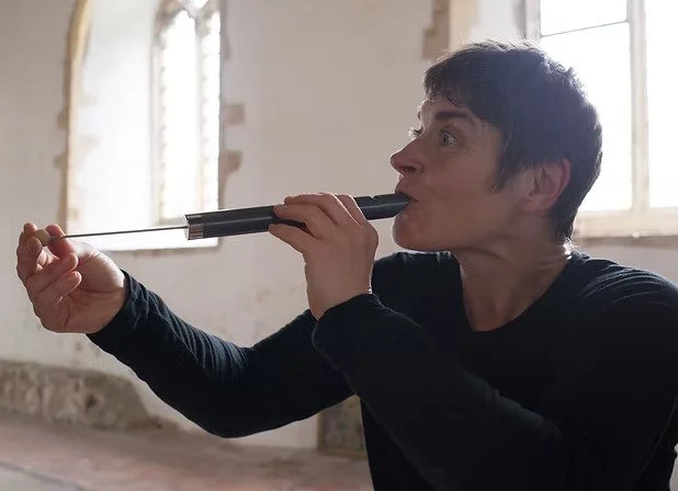 A woman with dark hair blowing into a pipe in an church with large windows.
