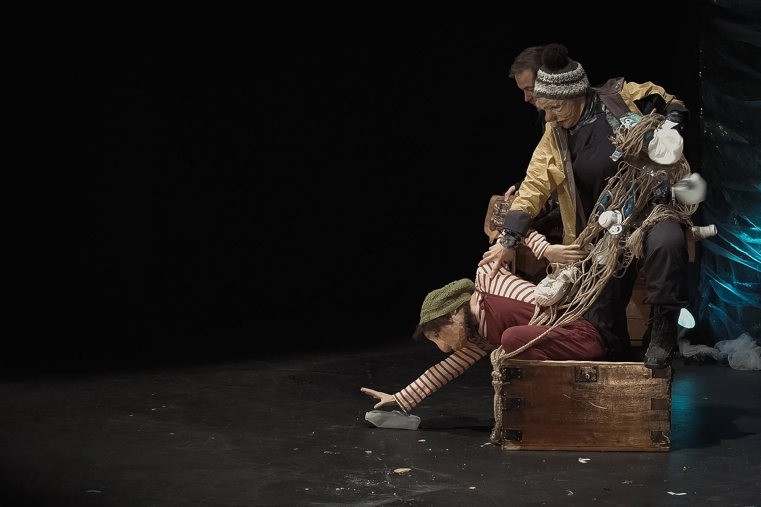 Theatrical performance showing two actors in elaborate costumes on a dark stage; one actor is reaching to pick up an object, while the other stands behind, with a suitcase and costume accessories.