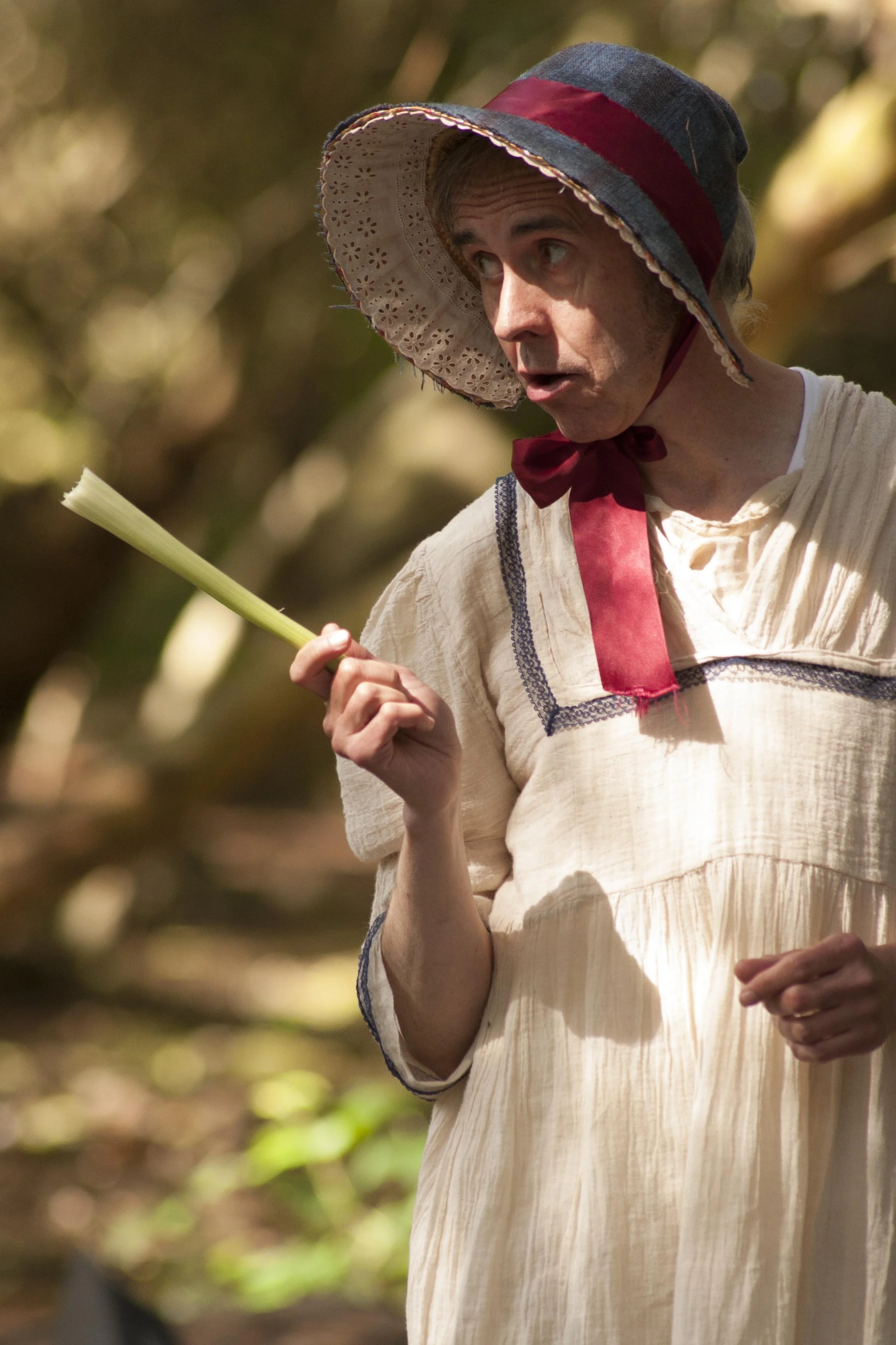 A woman in vintage clothing holding a leek, outdoors in a natural setting.