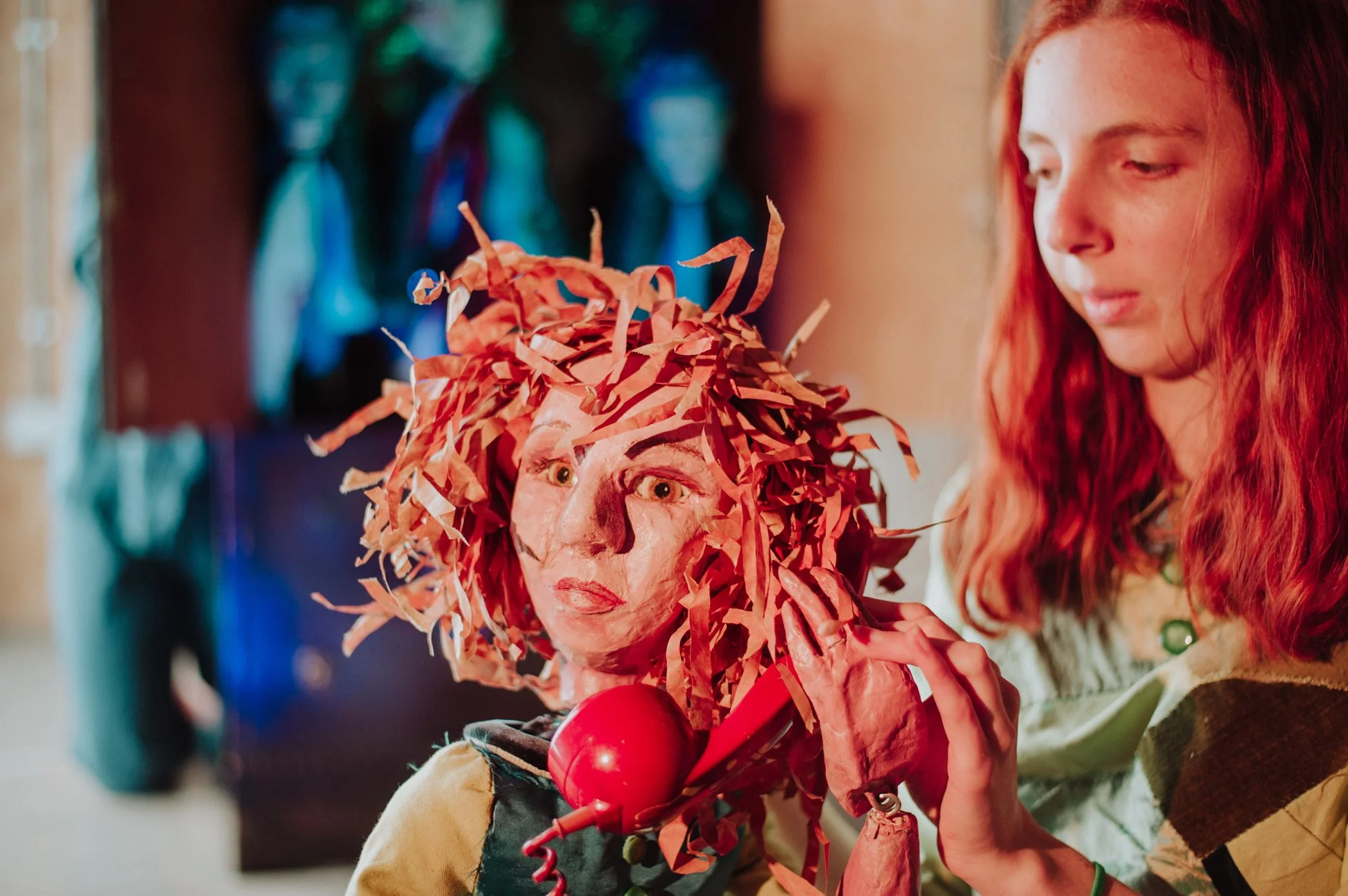 Young girl with red curly hair looking at an art puppet with orange paper curls, painted facial features, and a red hairdryer attached to the puppet.