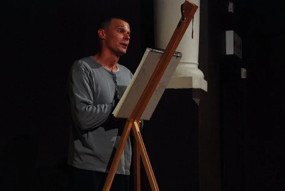 A man in a gray shirt standing next to an easel with a large sketchpad, speaking or performing on a dimly lit stage.