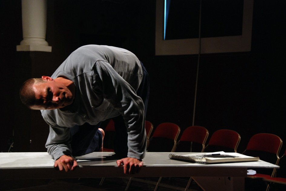 A man is crawling on a table in a dark room with red chairs around it, a notebook and papers on the table.
