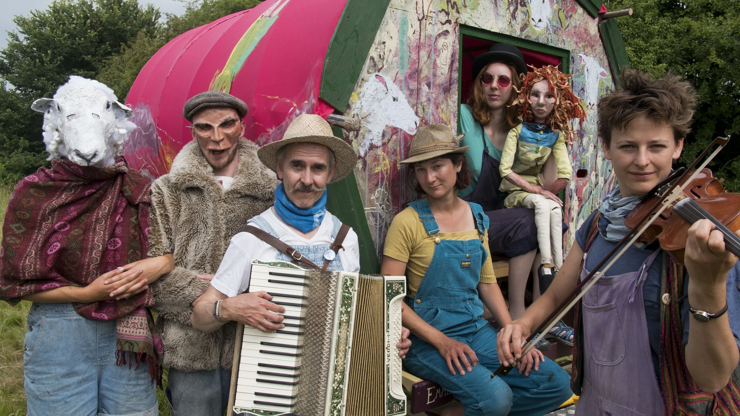 Group of eight people in colorful costumes and masks, some with musical instruments, gathered outside in front of a painted, whimsical trailer, with trees and greenery in the background.