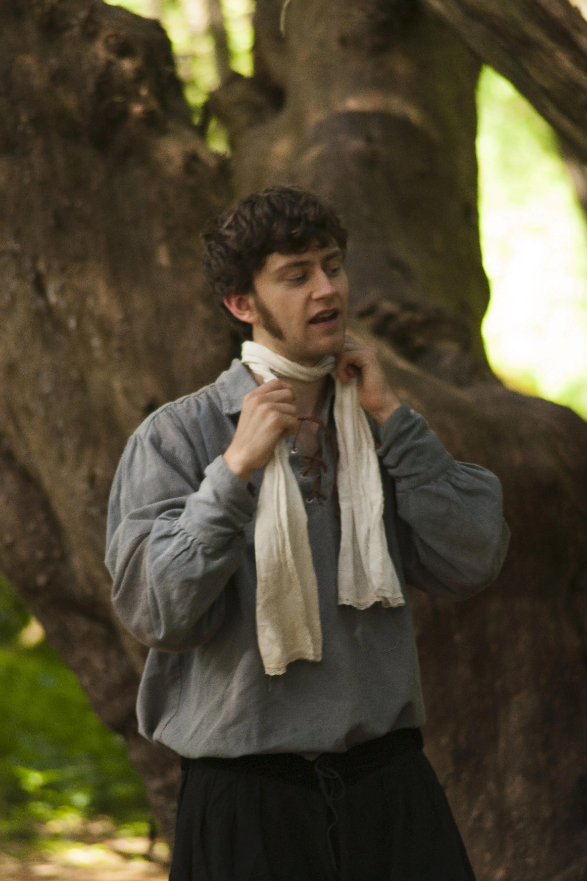 A young man adjusting a white scarf around his neck, standing outdoors in front of a large tree with green foliage.