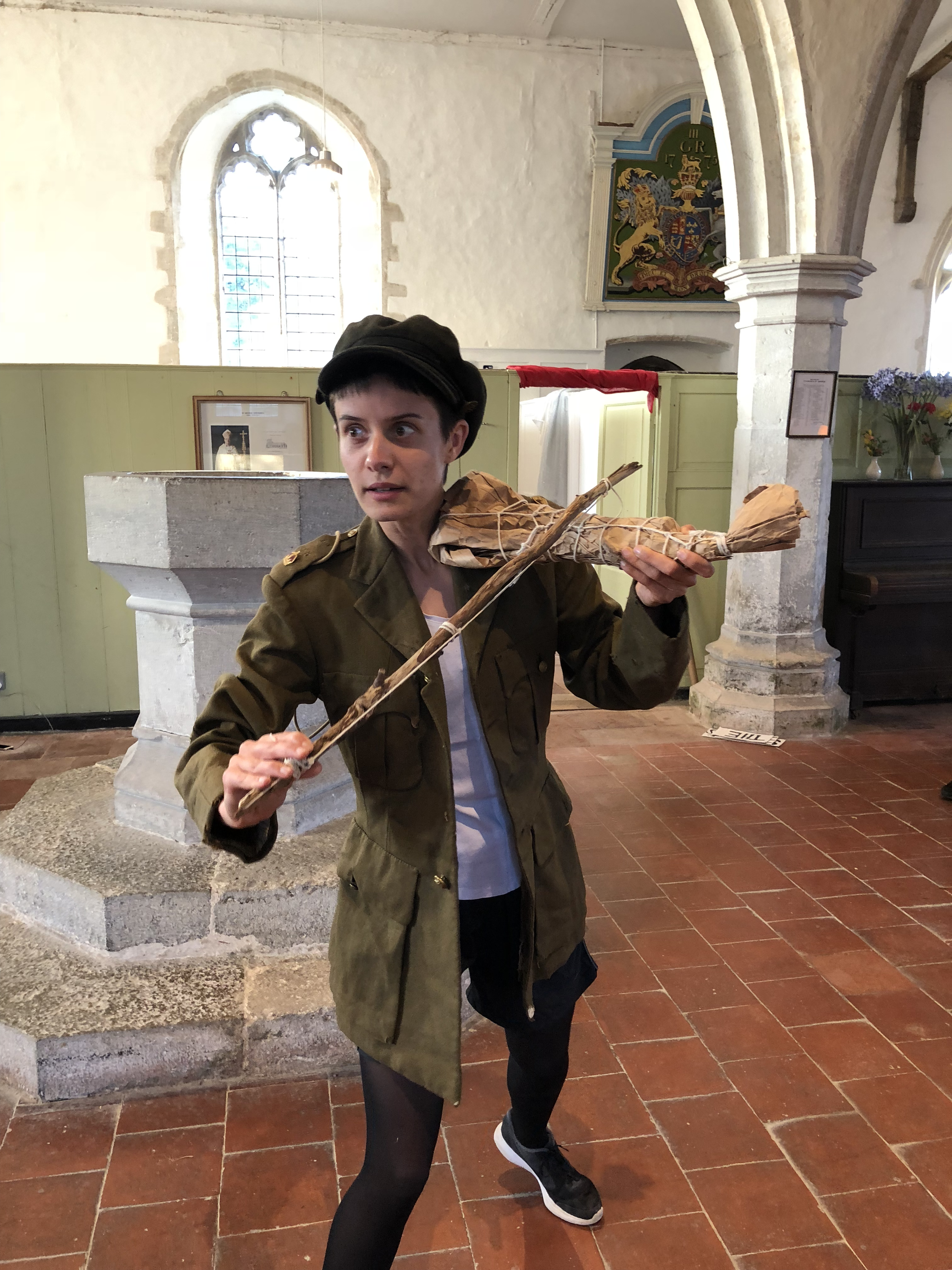 A person dressed as a soldier playing a violin, standing inside a church with a stained glass window in the wall behind.