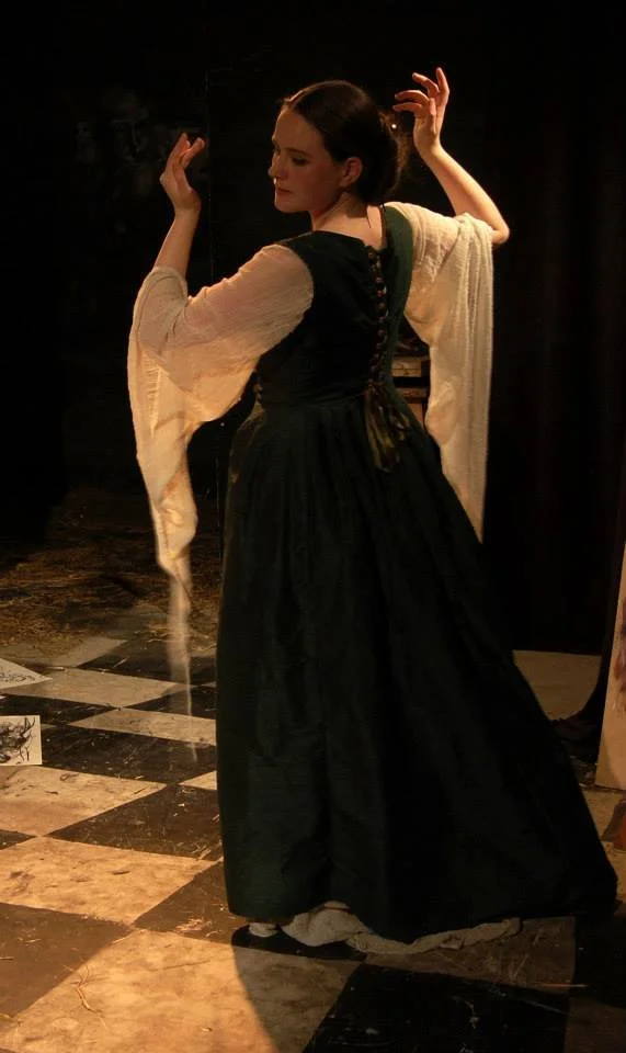 A woman in historical clothing, wearing a long black dress and a white waterfall sleeves, standing on a checkered floor and posing with her hands raised.