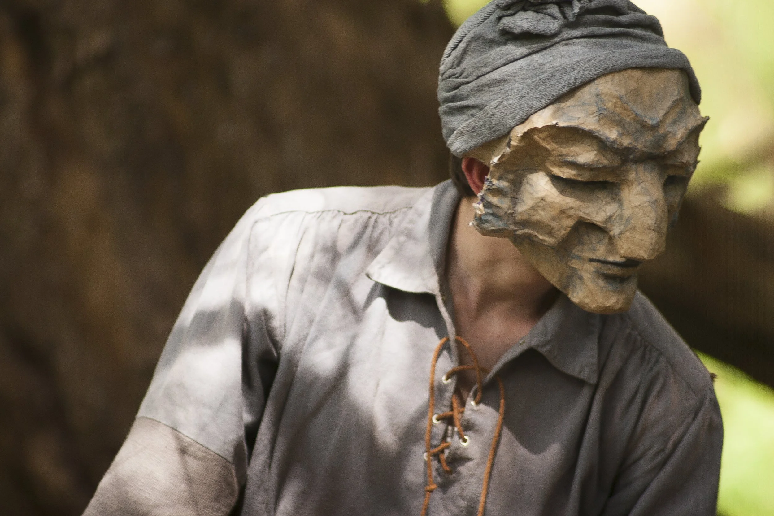A person wearing a gray hooded jacket and a gray headwrap or hat is seen with a mask made of carved wood or stone, shaped like a human face. The background appears natural, suggesting an outdoor setting.