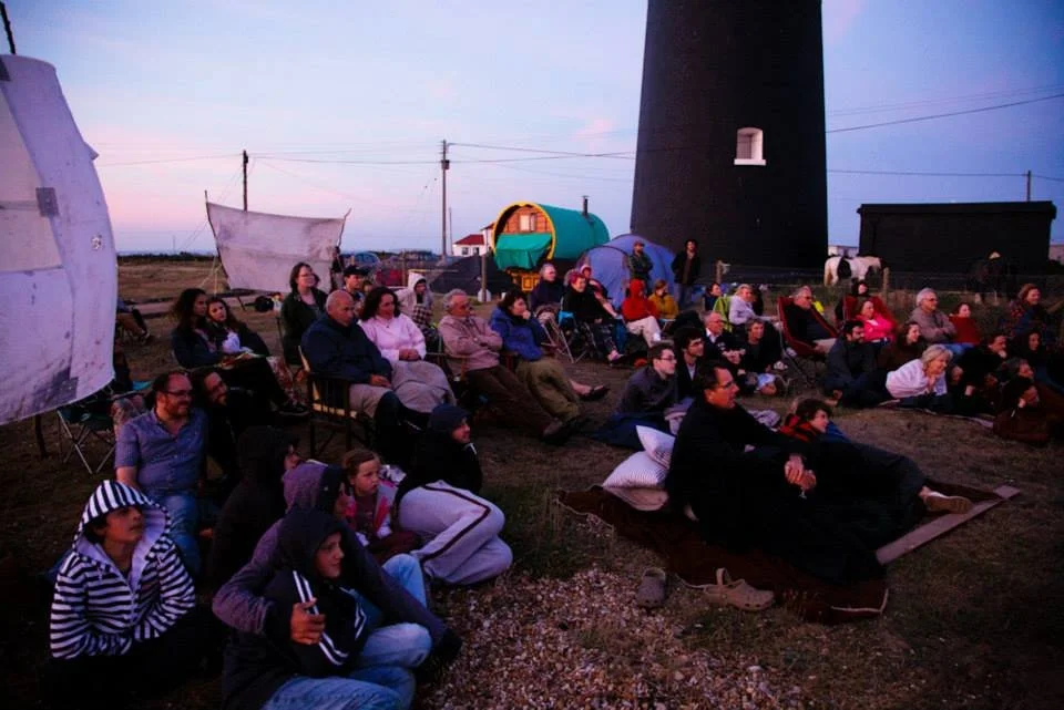 A group of people watching an outdoor movie under the evening sky near a lighthouse and tents.