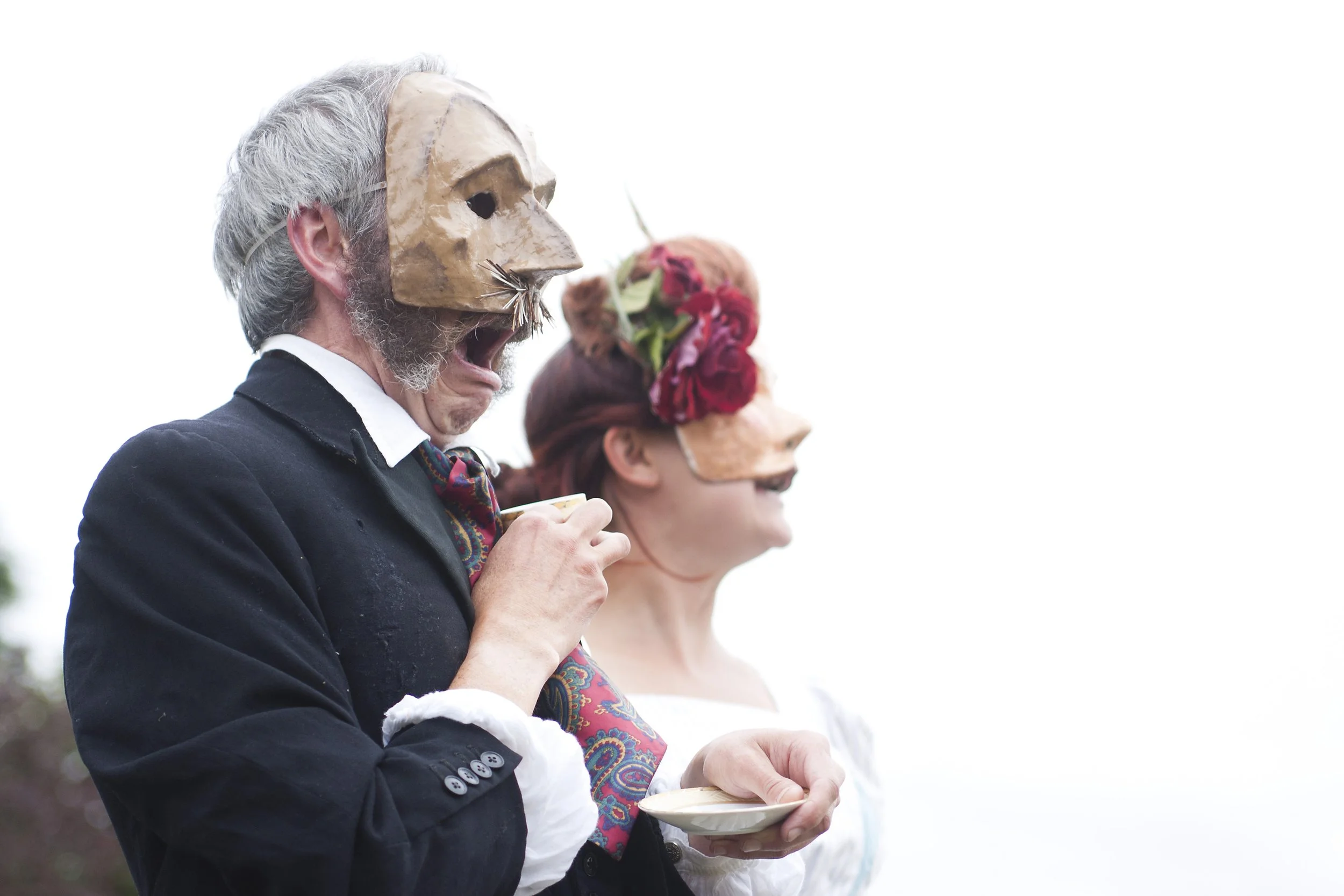 Two people wearing masks, one with a carved mask and the other with a colourful headpiece, stand outdoors holding cups.