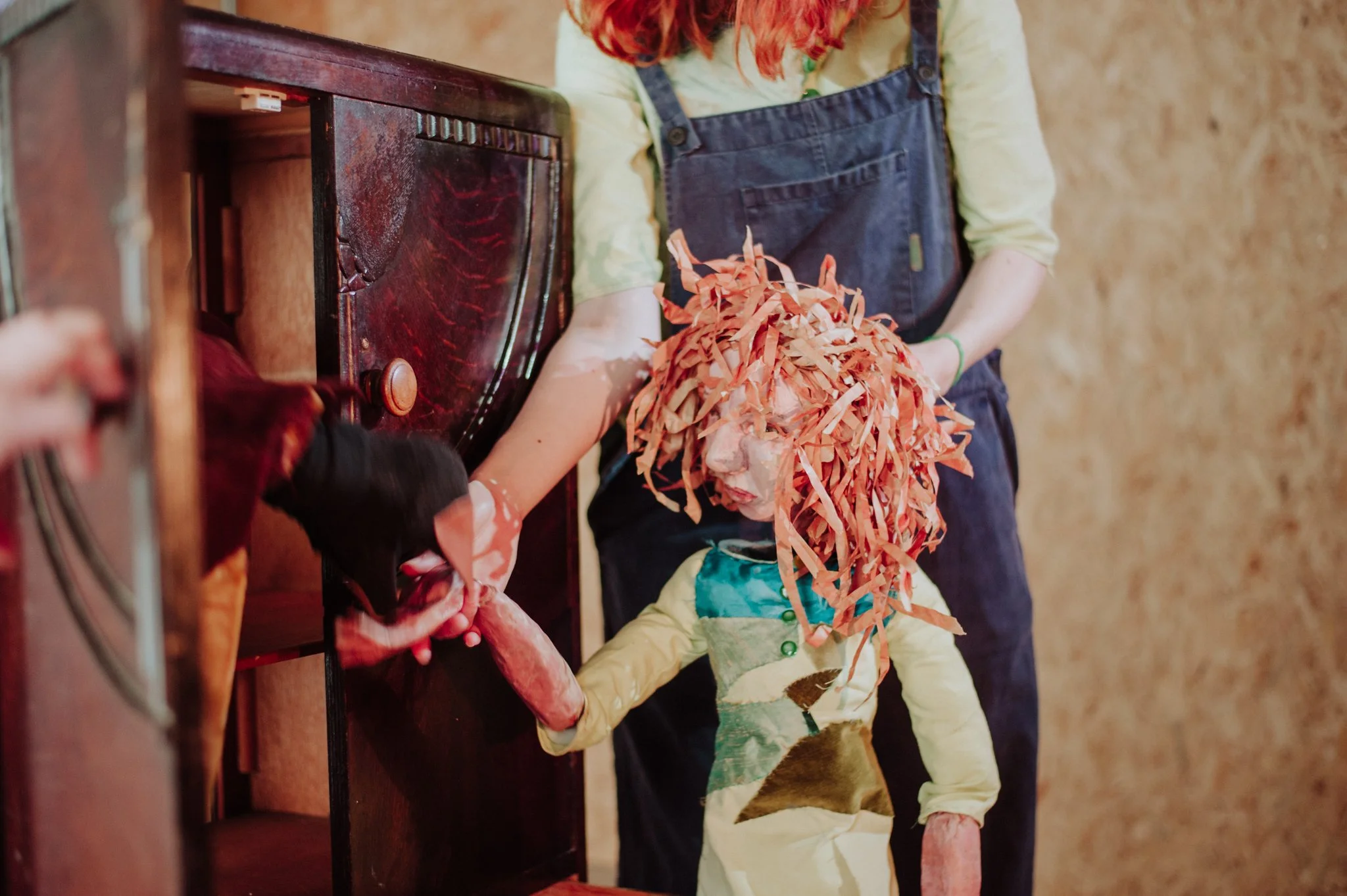 A puppet with a papier-mâché face and orange paper strips for hair, holding hands with a person, near a vintage wooden cabinet.