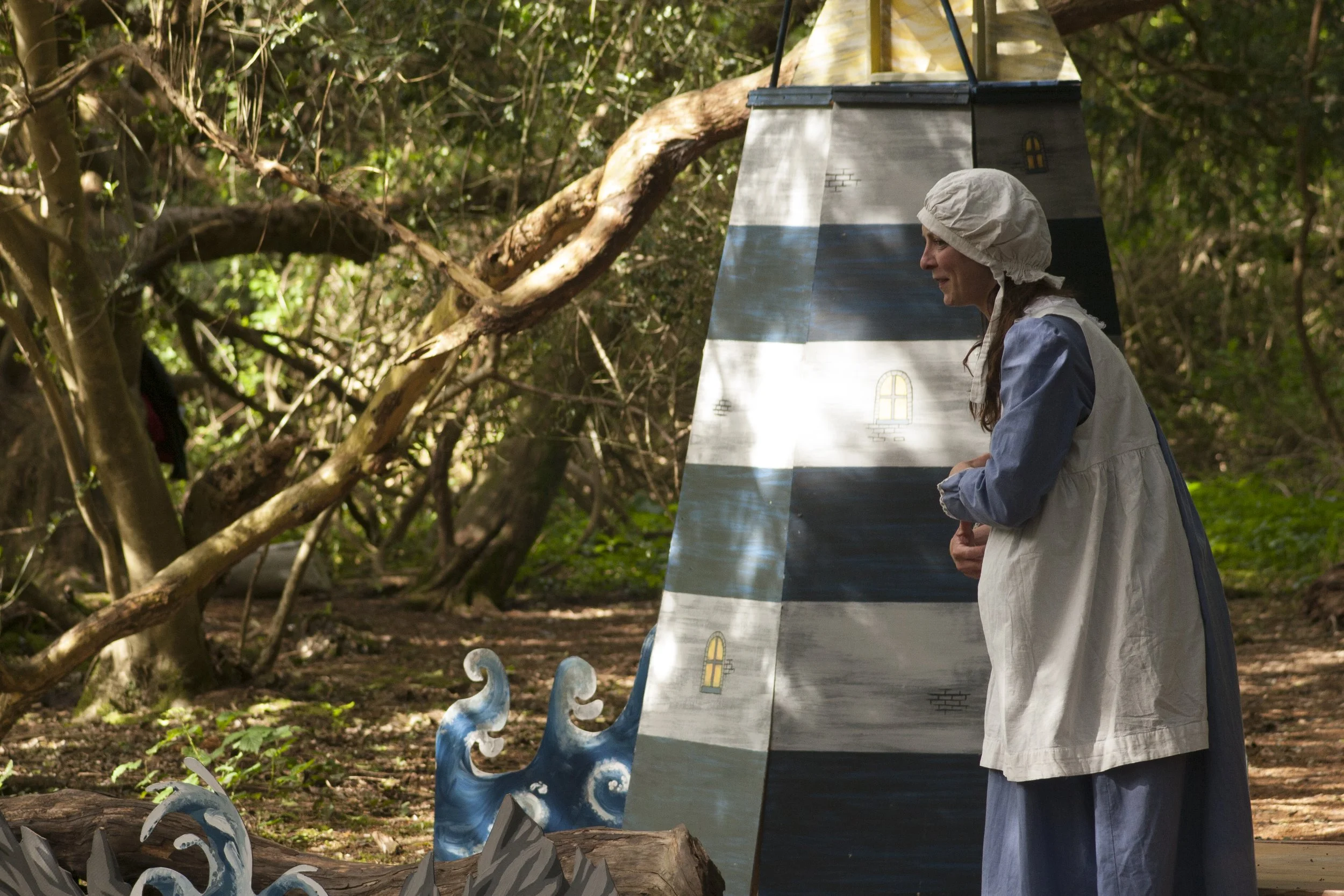 Woman dressed in a costume with a chimney design, standing outdoors in a wooded area near a painted prop resembling a castle tower.