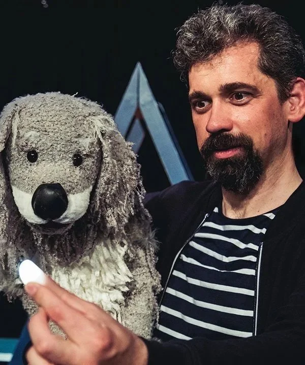 A man with curly hair and a beard holding a puppet dog. The man wears a black cardigan over a striped shirt, and the puppet resembles a dog with gray and white fur, black eyes, and a large black nose.