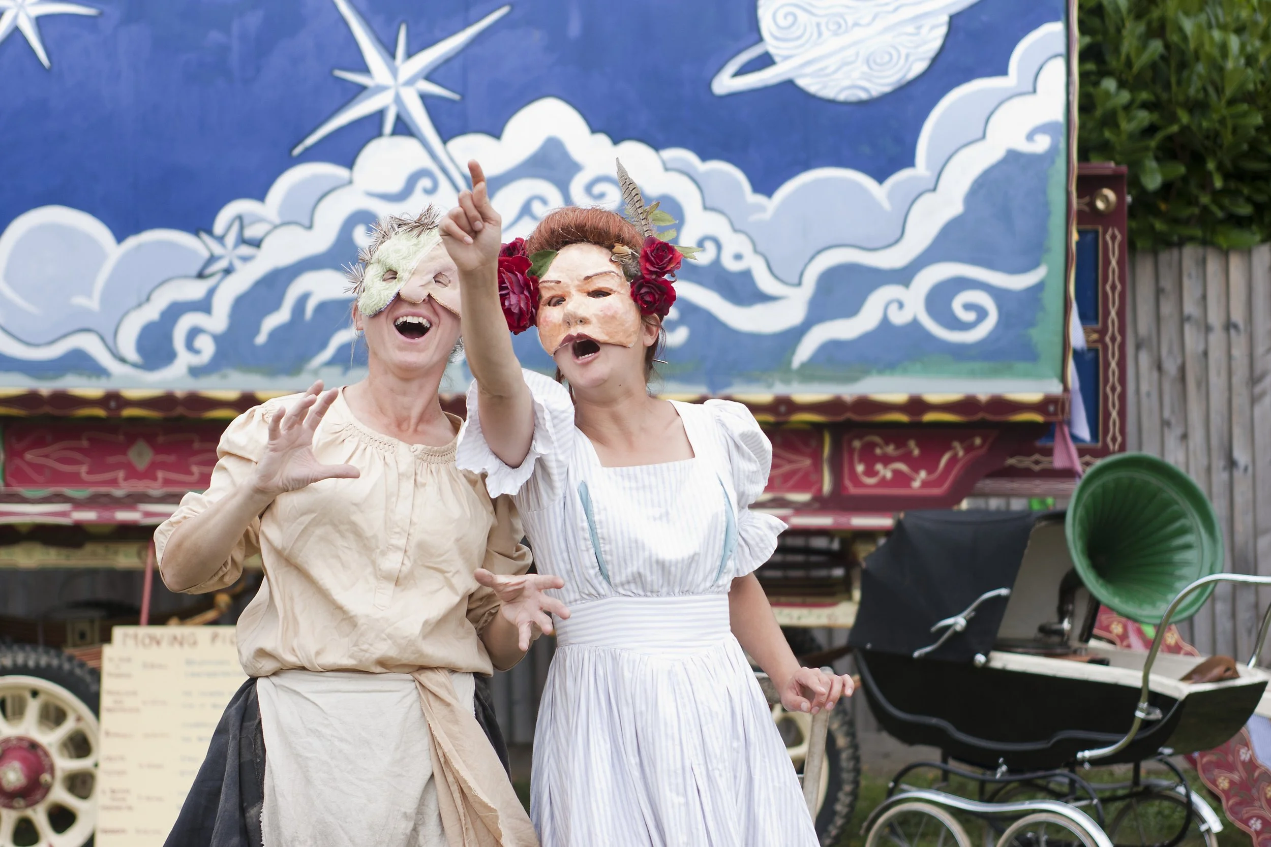 Two women dressed as theater characters with masks on their faces, one with a blonde wig and the other with red hair and flowers, standing in front of a colorful backdrop with clouds and a star, at an outdoor event.
