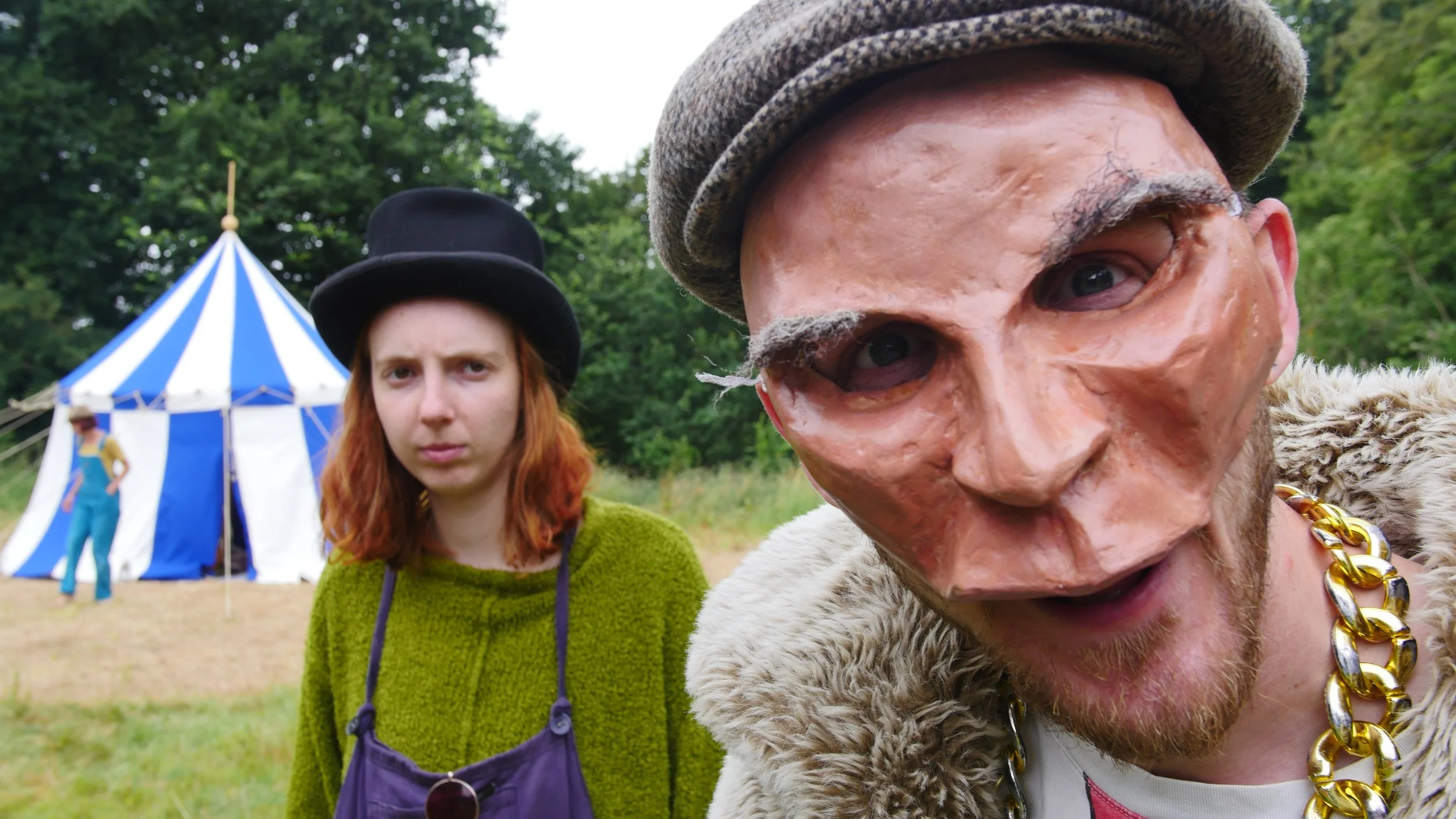 Two people standing outdoors during daytime, with a blue and white striped circus tent in the background. One person in the foreground wears a mask with exaggerated facial features, a beret, and a gold chain, while the other person in the background has red hair, a green sweater, and a black top hat.