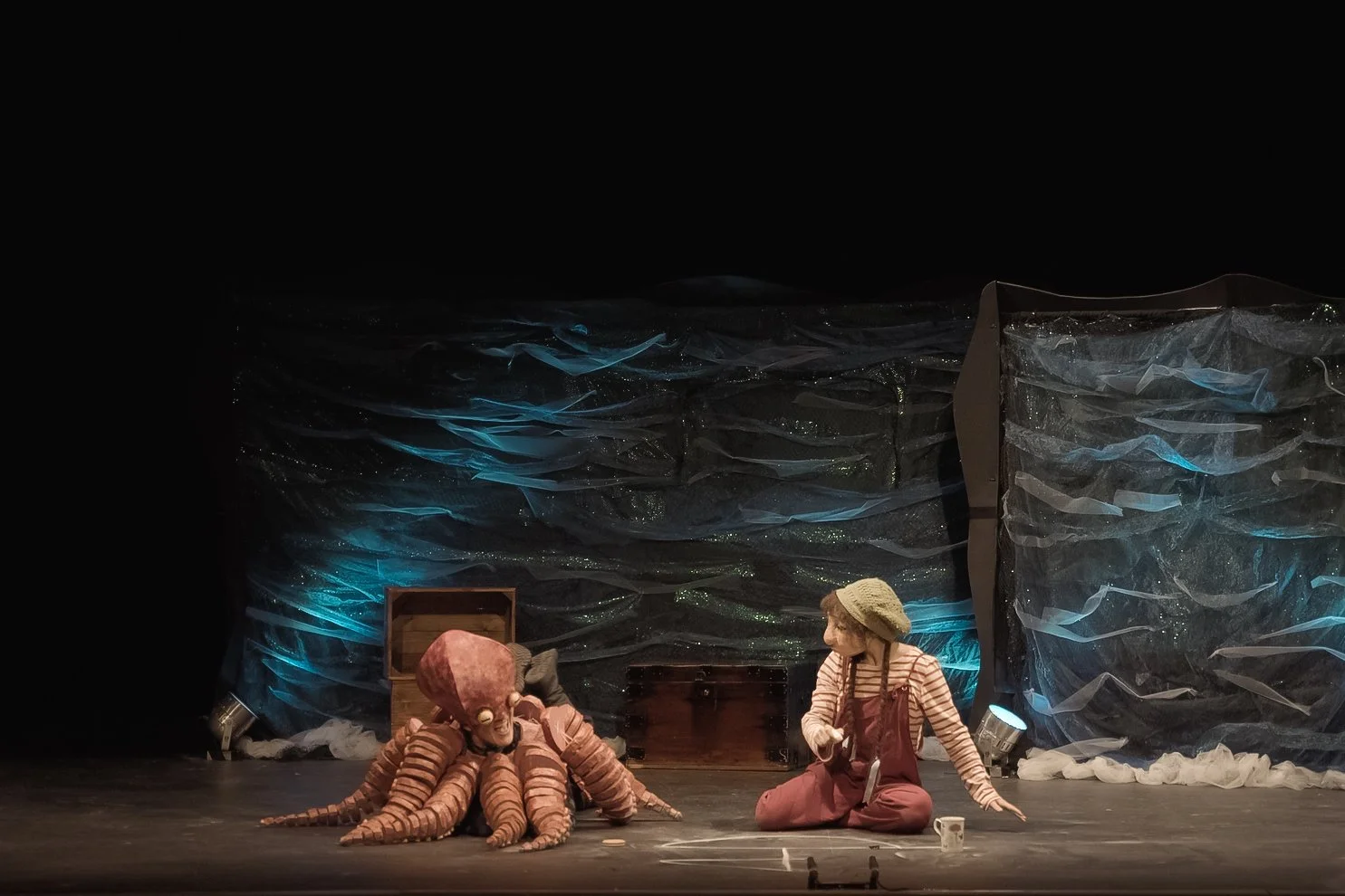 A stage scene from a theatrical play featuring two characters, one with an octopus-like costume and the other dressed as a girl with a straw hat, sitting on the floor amidst props and scenery resembling an underwater or ocean setting.