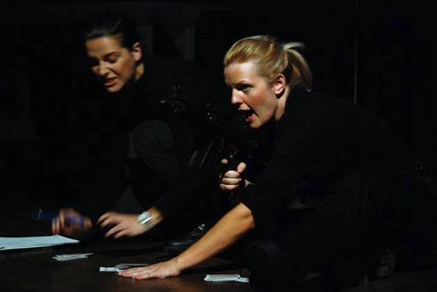 Two women are on the floor, one holding a gun and the other reaching for playing cards; both appear distressed or intense.